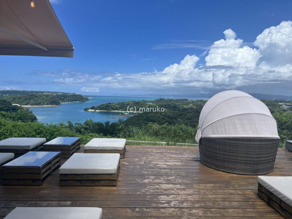 沖縄の高台カフェのテラスから見える青い海と緑の景色。白いソファとウッドデッキが並ぶ開放的な眺め。